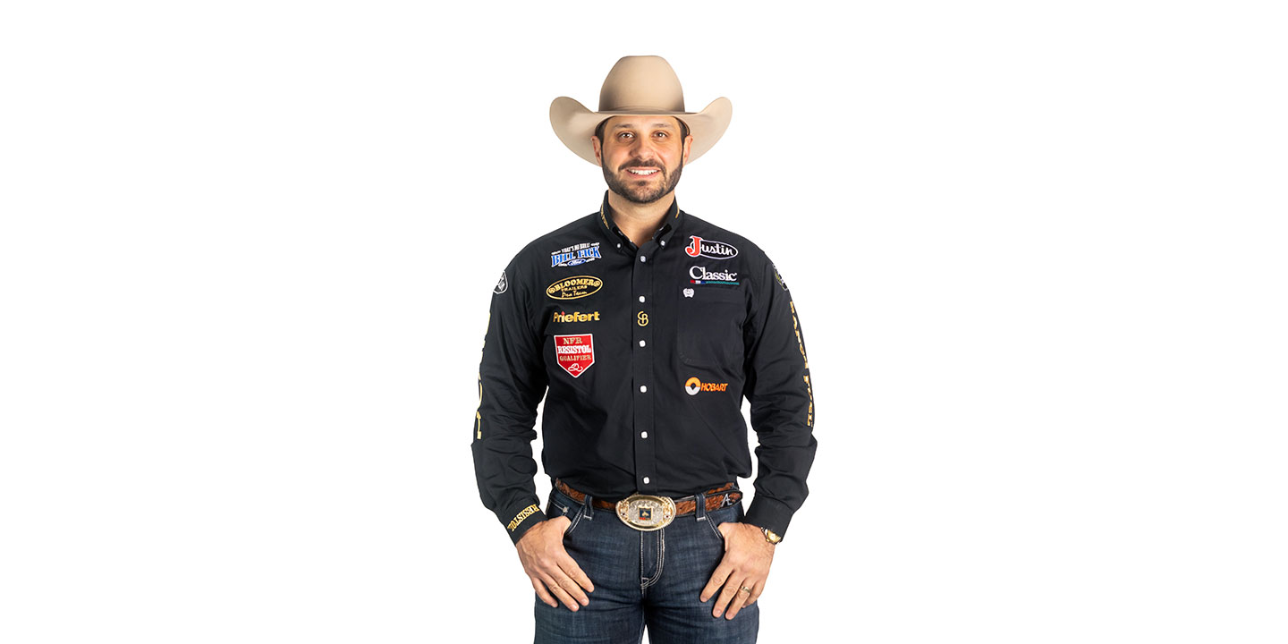 Anthony Lucia vistiendo una camisa negra y un sombrero de vaquero, sonriendo y posando frente a una pared blanca.